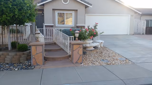 Front exterior view of a single-story house with a small staircase leading to the front door, a white railing, a tree, and a garden area with rocks and flowering plants. The house has a large white garage door and a concrete driveway.