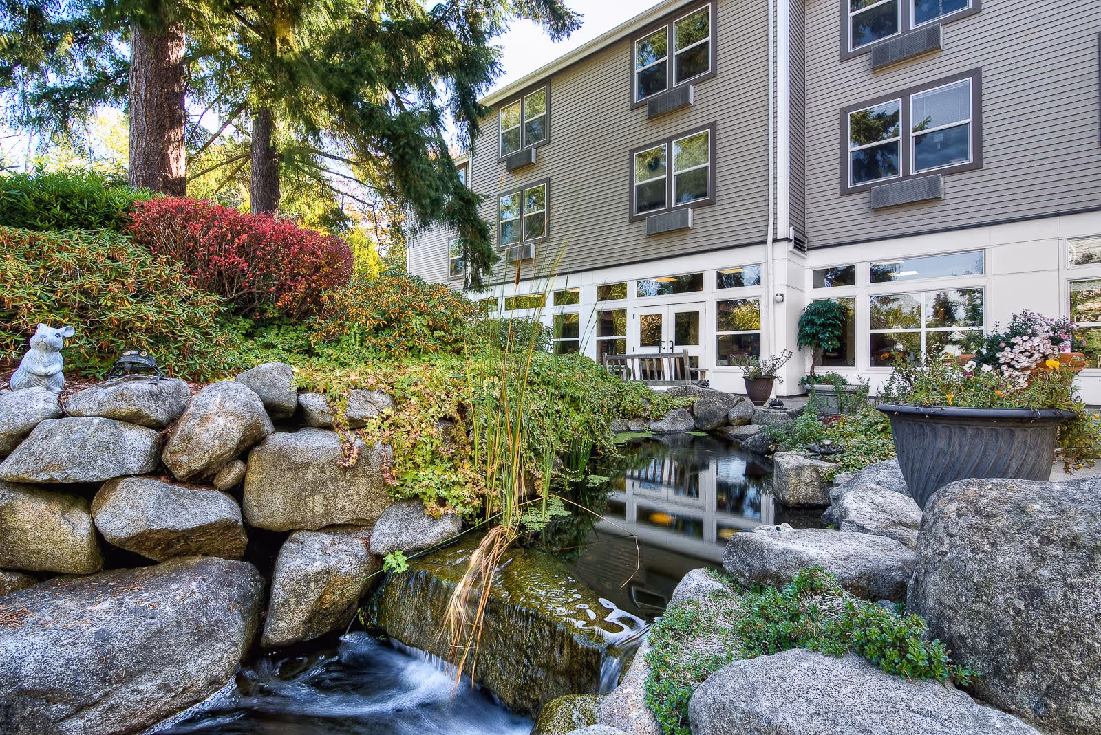 Outdoor garden area at GenCare Lifestyle Federal Way featuring a small pond with a waterfall surrounded by large rocks, lush greenery, colorful bushes, and potted plants. The background shows the exterior of a multi-story building with large windows reflecting the garden.