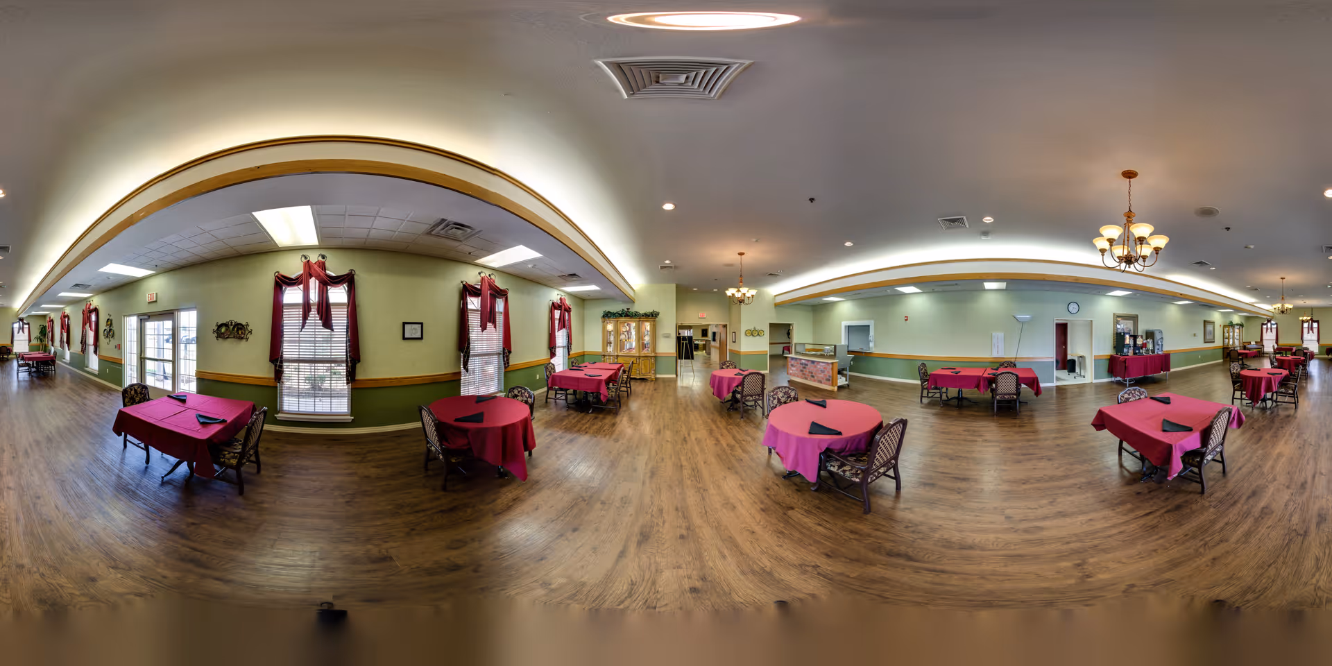 A spacious dining room with multiple tables covered in red tablecloths and black napkins. The room has wooden flooring, green walls with a wooden trim, windows with red curtains, and several chandeliers hanging from the ceiling. There is a cabinet with glass doors and a clock on the wall in the background.