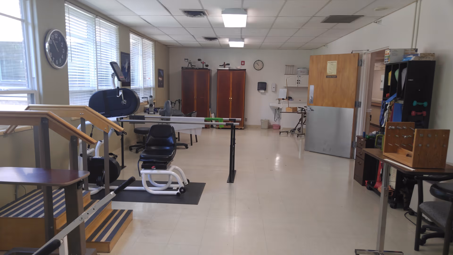 A rehabilitation or physical therapy room with exercise equipment including parallel bars, a seated exercise machine, and stairs with handrails. The room has large windows on the left side, a clock on the wall, wooden cabinets at the back, and various therapy tools and supplies on shelves and tables.