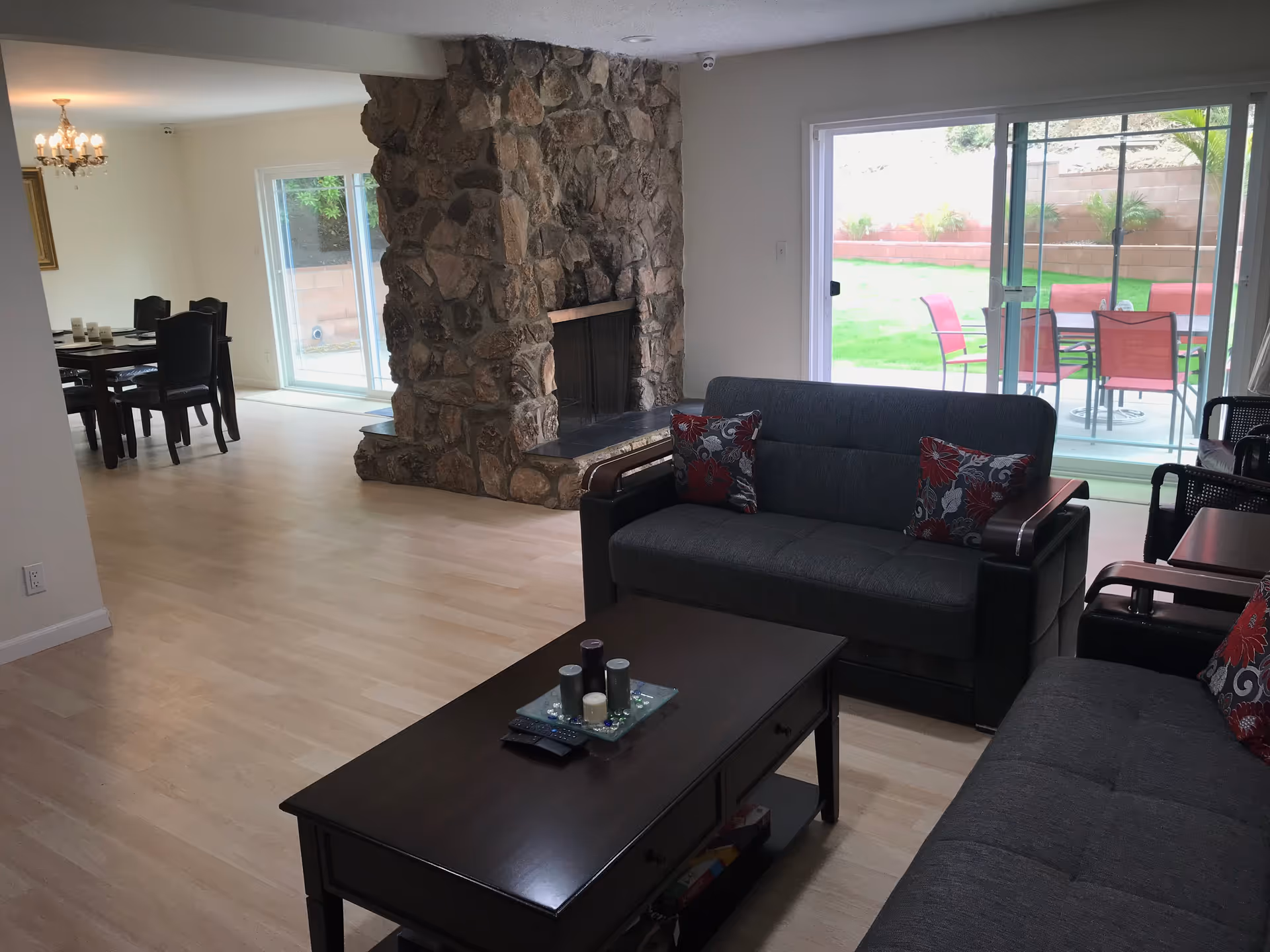 A living room with two dark gray sofas adorned with red and gray floral pillows, a dark wooden coffee table with candles and remote controls, and a large stone fireplace. In the background, there is a dining area with a dark wooden table and chairs, a chandelier, and sliding glass doors leading to an outdoor patio with red chairs and a table.