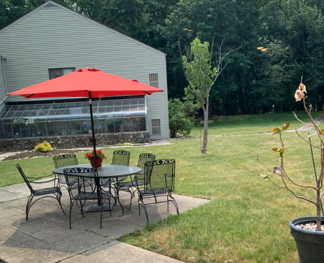 A patio with a metal table, six chairs and a red umbrella on a grassy lawn beside a light-colored building with a greenhouse and trees in the background.
