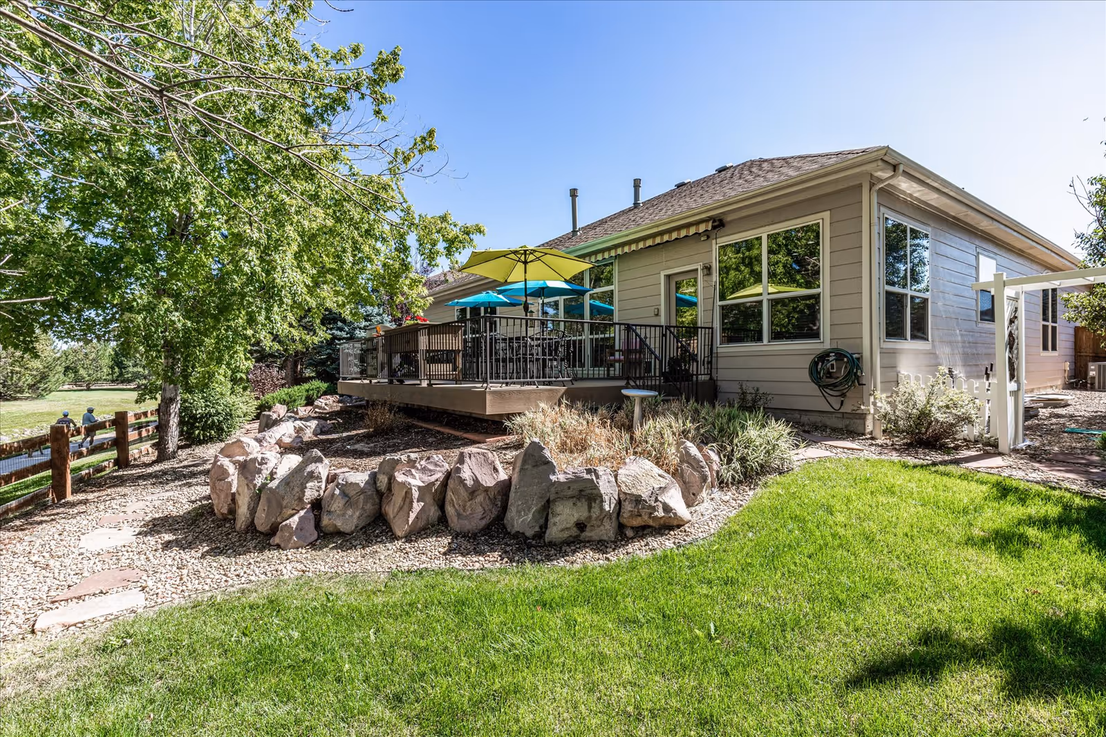 Outdoor view of a senior living facility showing a patio area with tables and umbrellas, surrounded by green grass, trees, rocks, and a wooden fence under a clear blue sky.