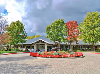 Front entrance of a single-story senior living facility with a circular driveway, landscaped flower island and trees under a partly cloudy sky.