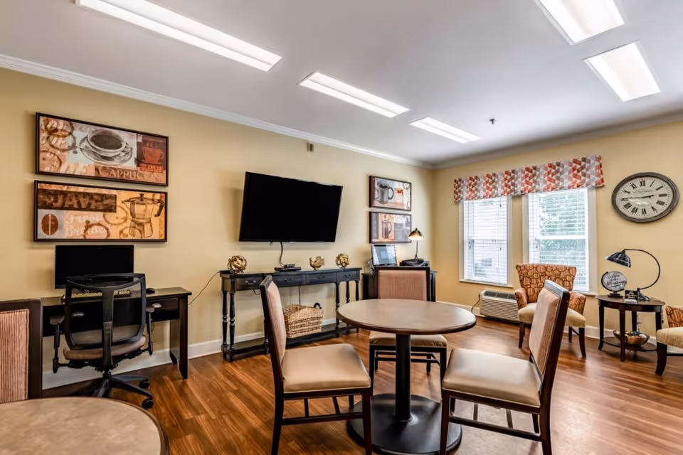 A well-lit common area with wooden flooring featuring a round table with four chairs in the center. There is a wall-mounted flat screen TV above a console table with decorative items. Two desks with chairs are positioned against the walls, and two windows with patterned valances allow natural light in. The room also includes two armchairs and a side table with a lamp and decorative globe, with a large wall clock nearby.