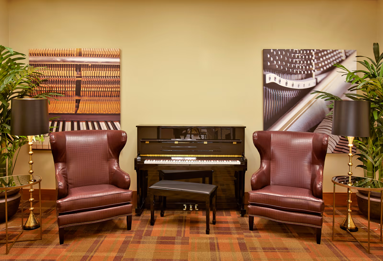 A cozy interior seating area featuring two maroon leather wingback chairs on either side of a black upright piano with a matching bench. Each chair is accompanied by a round side table with a gold base and a black lampshade. Two large framed close-up photographs of piano keys and strings hang on the beige wall behind the piano. There are also two tall green potted plants placed behind each chair. The floor is covered with a patterned carpet in warm tones.