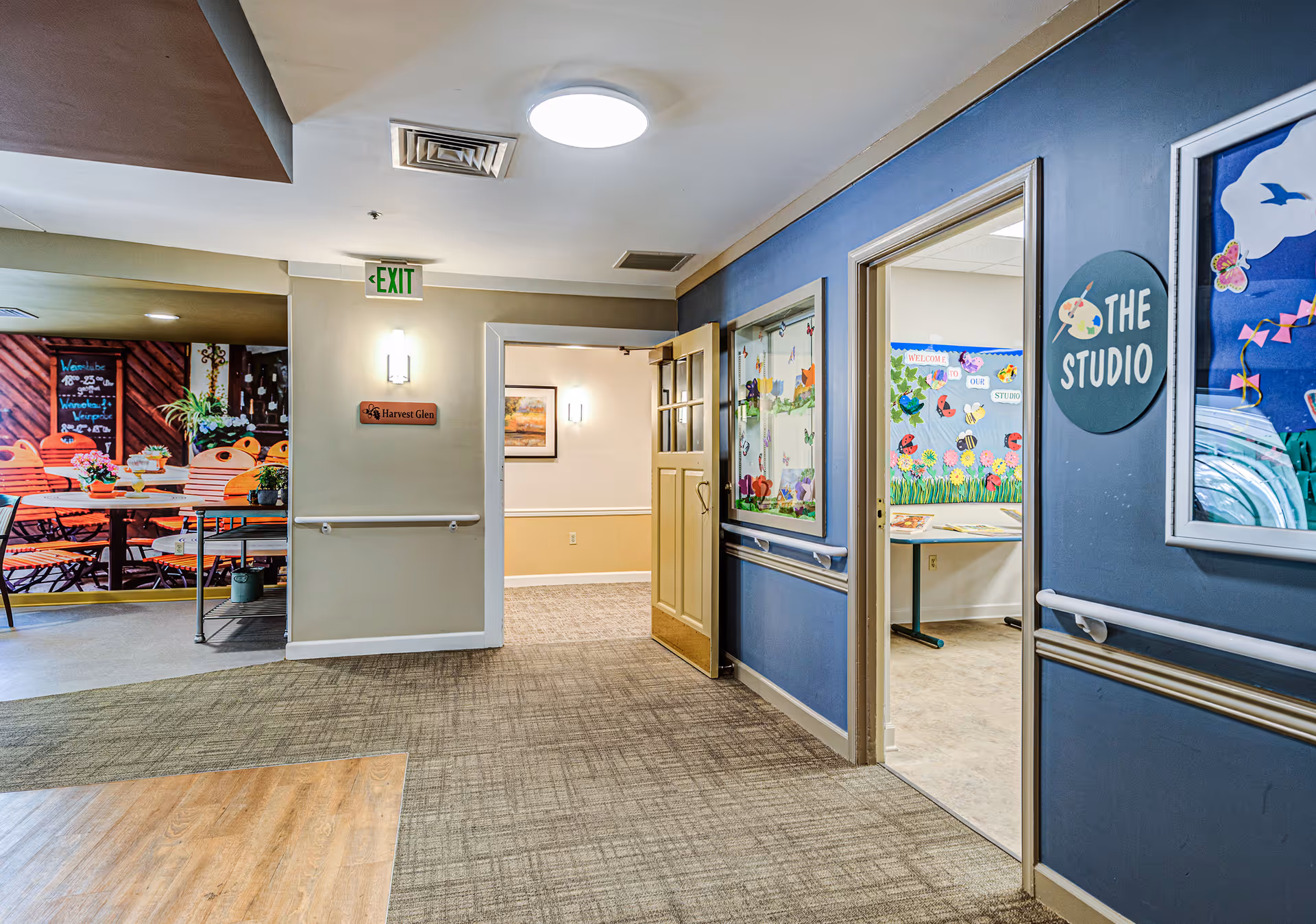 Interior hallway of a senior living facility with beige and blue walls, carpeted floor, and handrails. On the right, there is an open door leading to a room labeled 'The Studio' with colorful decorations on the walls. On the left, there is a wall mural depicting an outdoor café scene with tables and chairs. An exit sign is visible above a door in the center background.