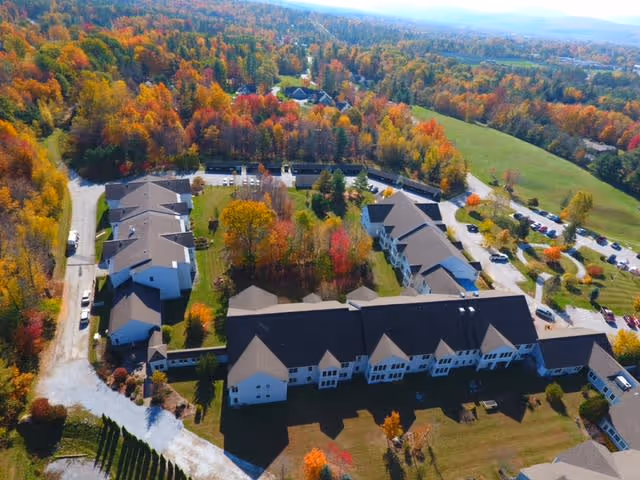 Aerial view of a senior living facility named Gables at East Mountain surrounded by colorful autumn trees and greenery, with multiple buildings, parking areas, and open fields visible.