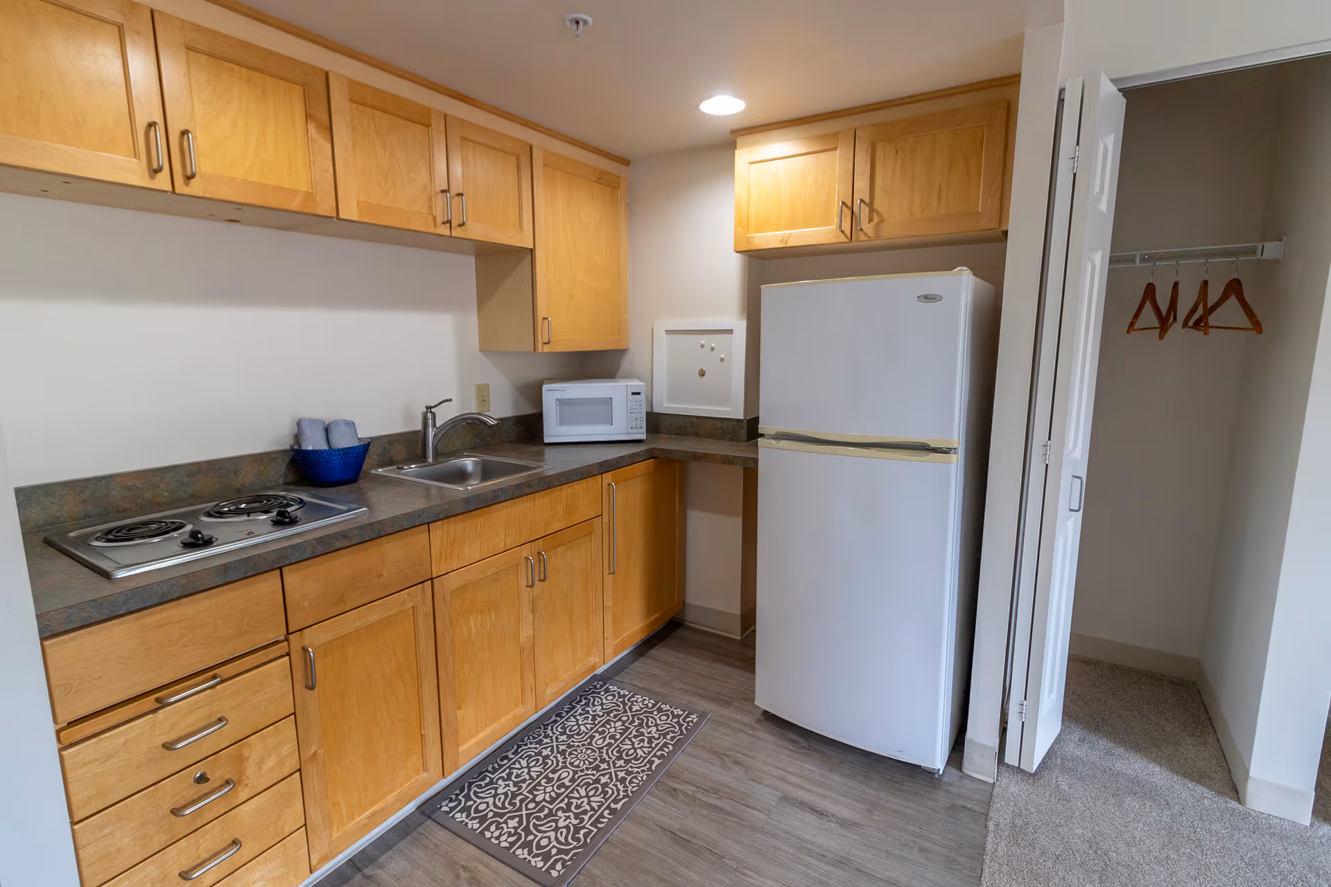 A small kitchen area with wooden cabinets, a white refrigerator, a microwave, a sink, and a two-burner electric stove. There is a patterned floor mat in front of the sink and stove. To the right, there is an open closet with a few wooden hangers inside.