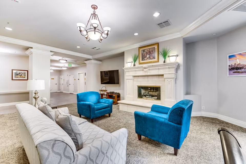 A cozy senior living facility common area with a white patterned sofa and two bright blue armchairs arranged around a stone fireplace. Above the fireplace is a framed painting and two potted plants. A flat-screen TV is mounted on the wall to the right. The room has light gray walls, carpeted flooring, and ceiling lights including a chandelier. There are framed pictures on the walls and a hallway visible in the background.