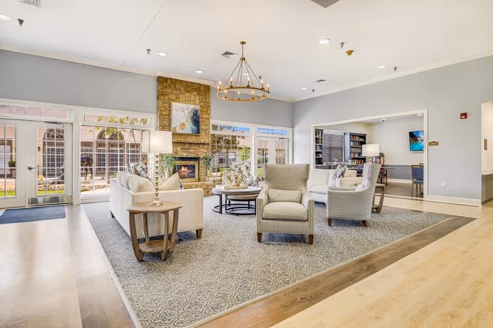 A bright and spacious senior living facility common area with comfortable seating including armchairs and sofas arranged around a stone fireplace. Large windows and glass doors let in natural light, and a modern chandelier hangs from the ceiling. The room features light-colored walls, a patterned area rug, and wooden flooring. In the background, there is an adjoining room with bookshelves and additional seating.