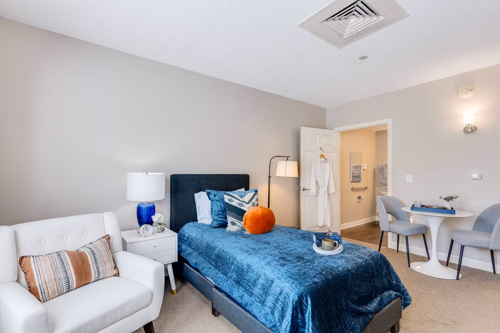 A cozy bedroom in a senior living facility featuring a single bed with a dark blue headboard and blue bedding, accented with multiple pillows including a round orange one. Next to the bed is a white nightstand with a blue lamp, a small clock, and a flower vase. A white armchair with a patterned cushion is positioned nearby. In the background, there is a small round white table with two gray chairs. The door to an adjacent bathroom is open, showing towels and bathroom fixtures. The room has light-colored walls and carpeted flooring.