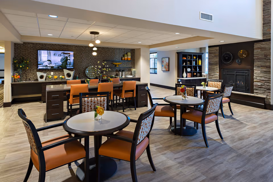 Communal dining and lounge area with round tables and orange chairs, a counter with bar stools, a wall-mounted TV and a fireplace.