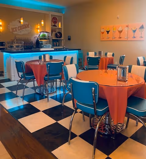 A retro-style dining area with round tables covered in orange tablecloths and blue and white chairs. The floor has a black and white checkered pattern. In the background, there is a counter with a glowing blue light underneath and an ice cream sign on the wall. The walls are decorated with paintings of various ice cream desserts.