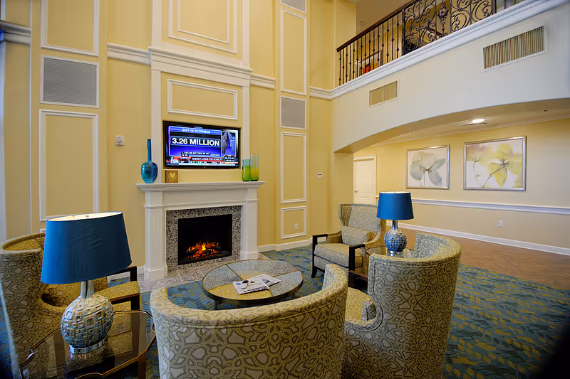 A spacious and elegant senior living facility common area with high ceilings, a fireplace, and a mounted flat-screen TV above it. The room features patterned armchairs arranged around a round glass coffee table with magazines, two blue table lamps on side tables, and framed floral artwork on the walls. A balcony with decorative iron railings overlooks the area.