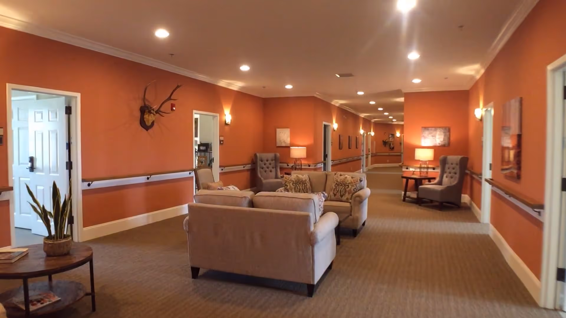 A long hallway in a senior living facility with orange walls and beige carpet. The hallway features comfortable seating areas with beige sofas and armchairs, side tables with lamps, wall-mounted handrails, and decorative wall art. There is a mounted deer head on the wall and several open doorways leading to other rooms.