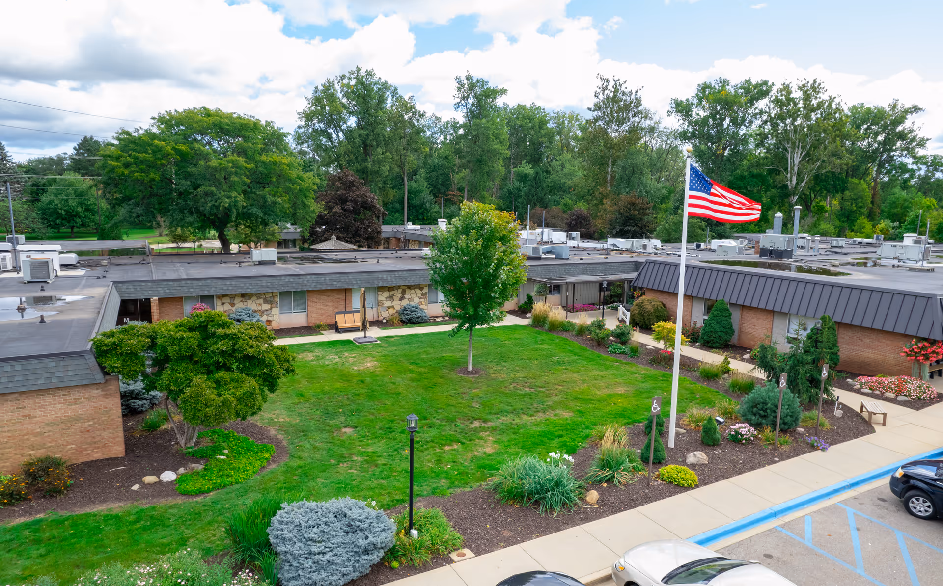 Aerial view of a single-story care facility courtyard with a central lawn, landscaped beds, an American flagpole, and adjacent parking.