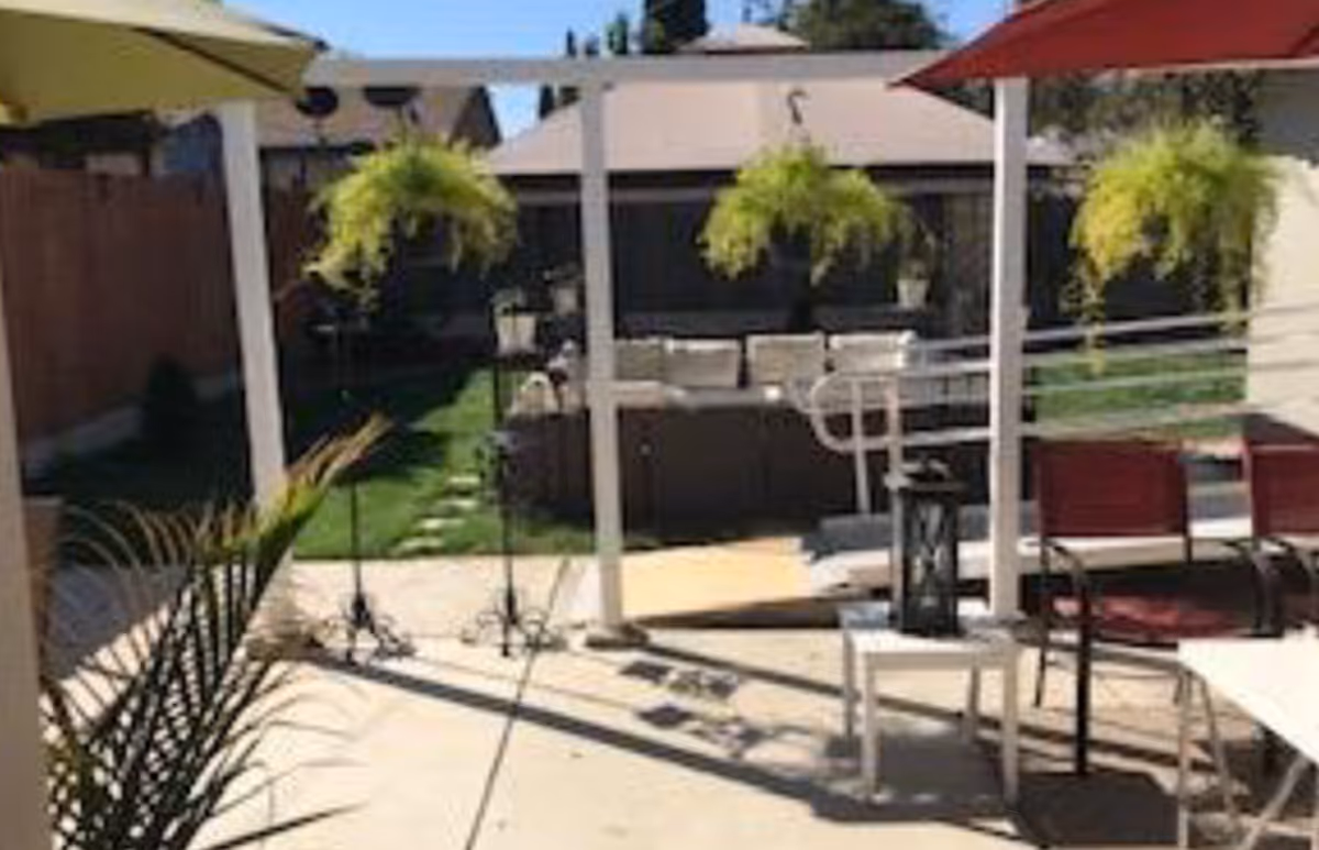 Outdoor patio area with hanging plants, patio furniture including chairs and a table, and a small garden with grass and stepping stones in the background.