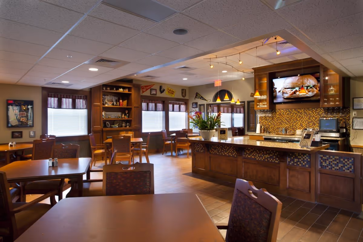 A cozy dining area in a senior living facility featuring wooden tables and chairs, a central counter with a flower arrangement, pendant lighting, and a wall-mounted TV displaying a sandwich. The room has large windows with blinds and decorative items on shelves and walls.