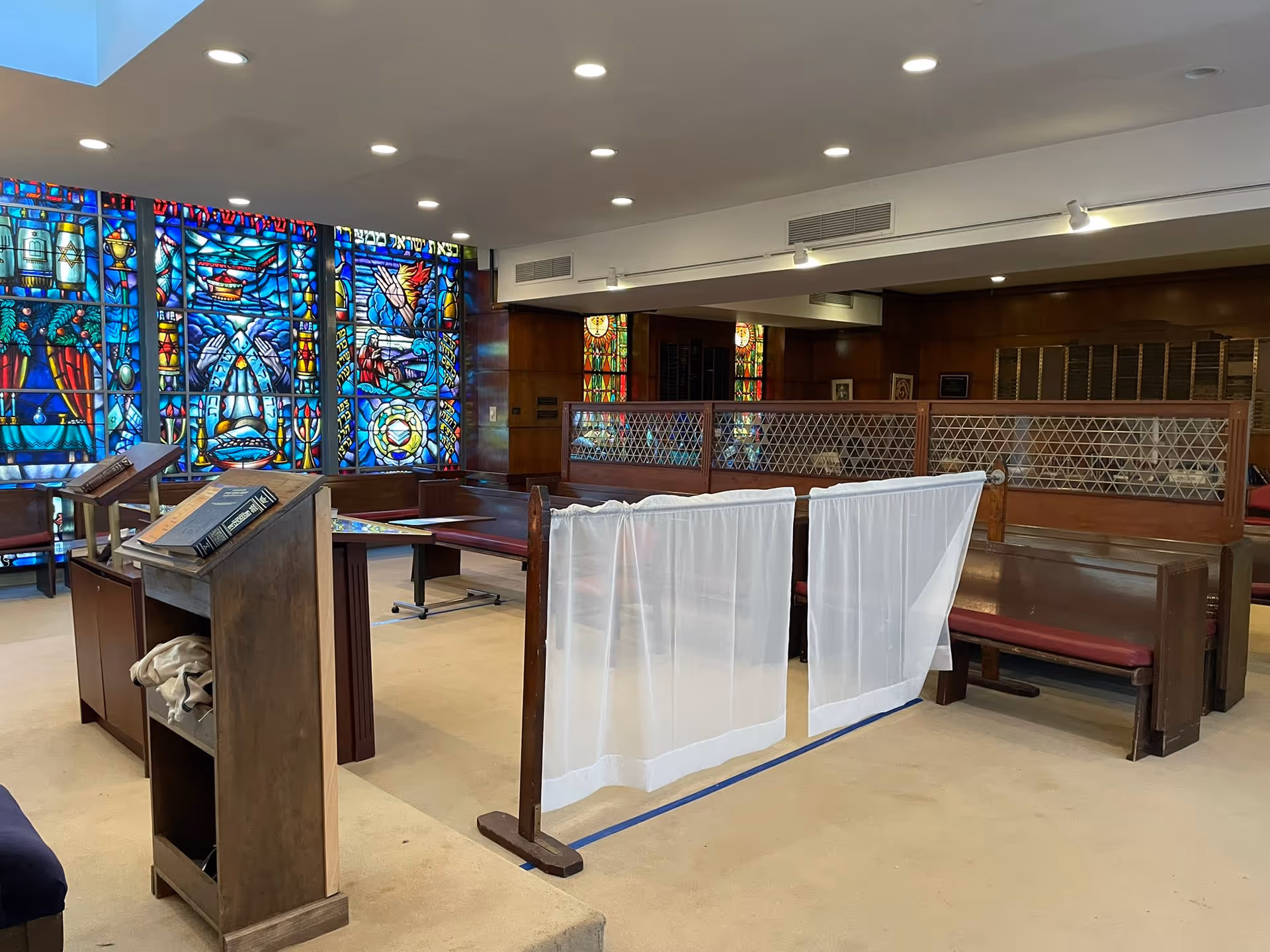 Interior of a room with wooden pews and lecterns, featuring large colorful stained glass windows along one wall. The room has a beige carpet and white ceiling with recessed lighting. A wooden divider with white fabric panels separates part of the seating area.