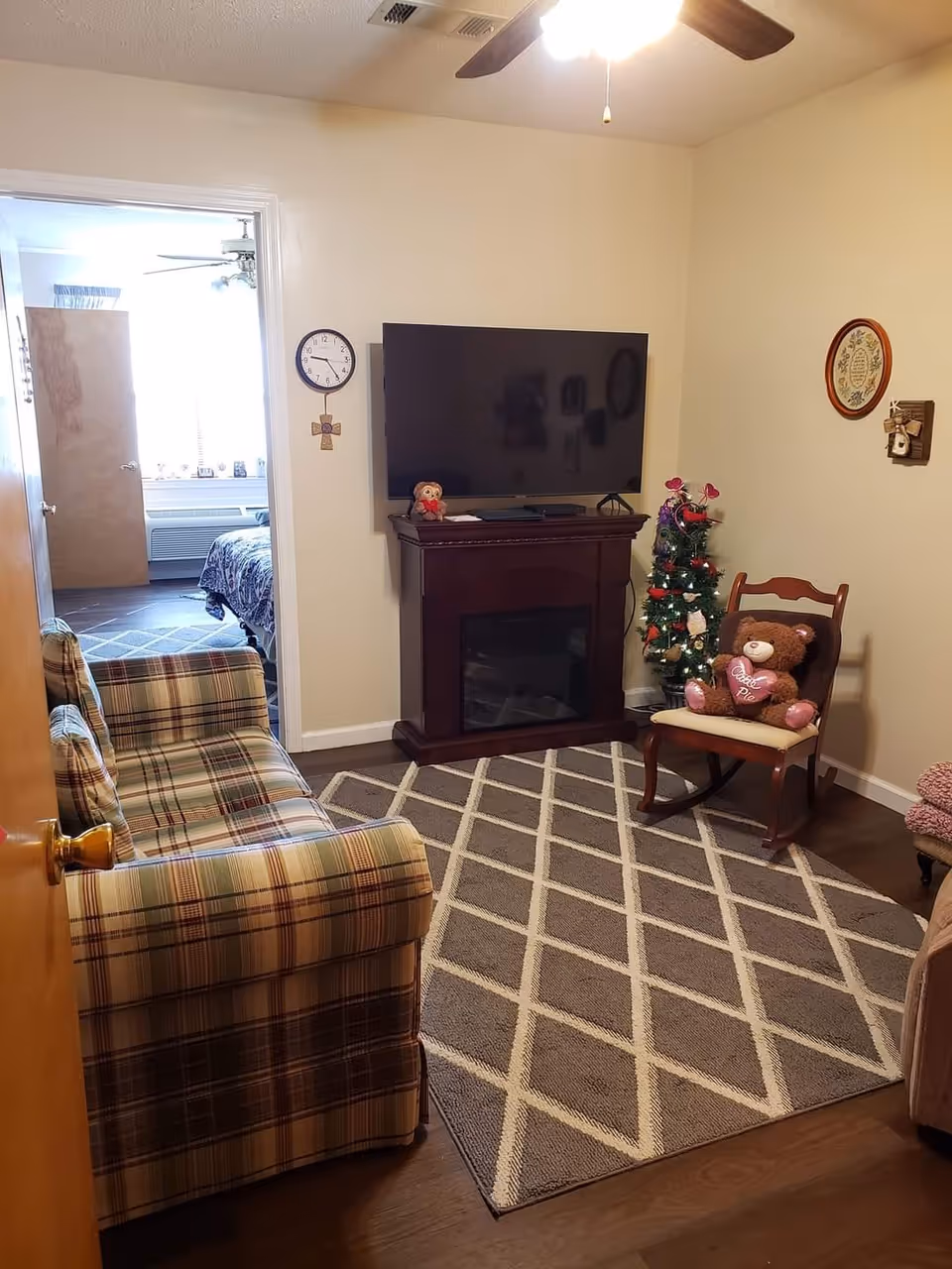 A cozy living room with a plaid sofa on the left, a patterned area rug on the wooden floor, and a flat-screen TV mounted above a dark wooden electric fireplace. A small decorated Christmas tree and a wooden rocking chair with a teddy bear holding a heart pillow are on the right. A doorway leads to a bedroom with a bed and a ceiling fan visible.