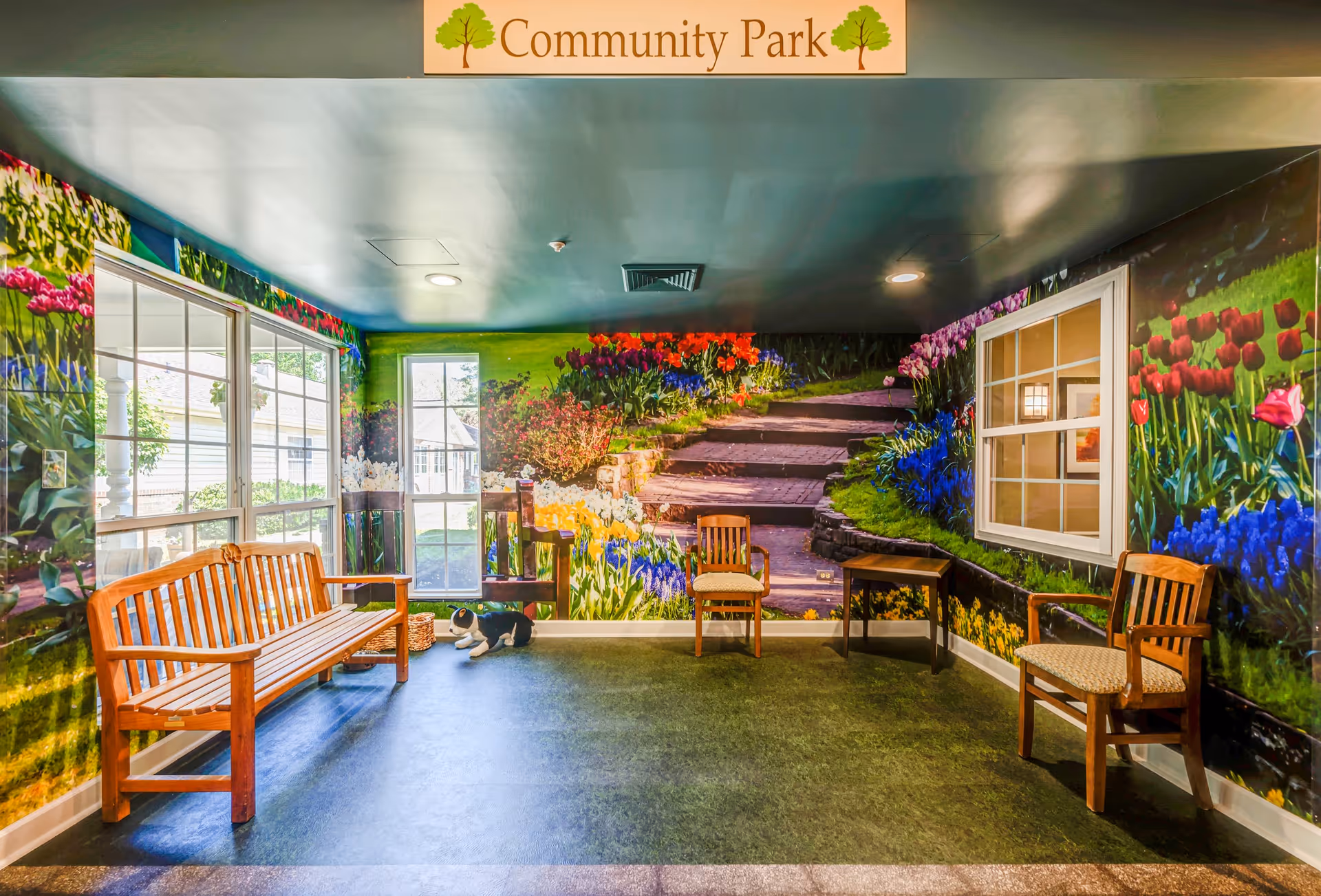 An indoor seating area with wooden benches and chairs arranged around the room. The walls are decorated with a large mural depicting a colorful garden with flowers and a stone pathway. There are windows on two walls, and a sign above reads 'Community Park'.