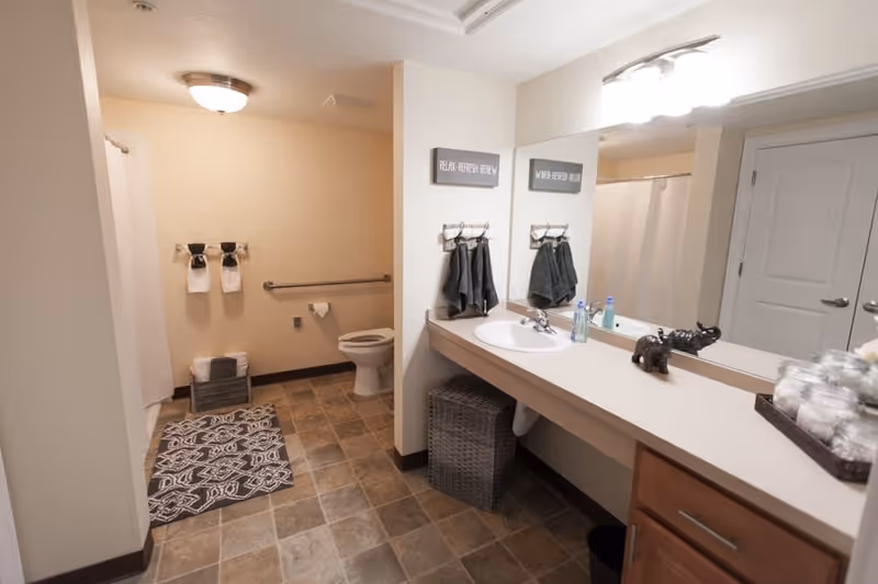 A clean and spacious bathroom with a toilet, a shower with a white curtain, a large mirror above a countertop with a sink, and neatly arranged towels. The floor has patterned tiles and there are decorative signs on the wall.
