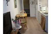 Narrow carpeted senior-living apartment interior with a small kitchenette on the right, a round dining table with two chairs, a TV and a vase of flowers.