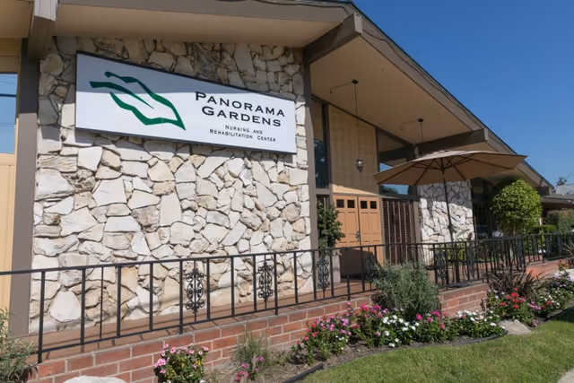 Exterior view of Panorama Gardens Nursing and Rehabilitation Center showing a stone facade with a sign, a railing, a garden with flowers, and a large umbrella providing shade near the entrance.