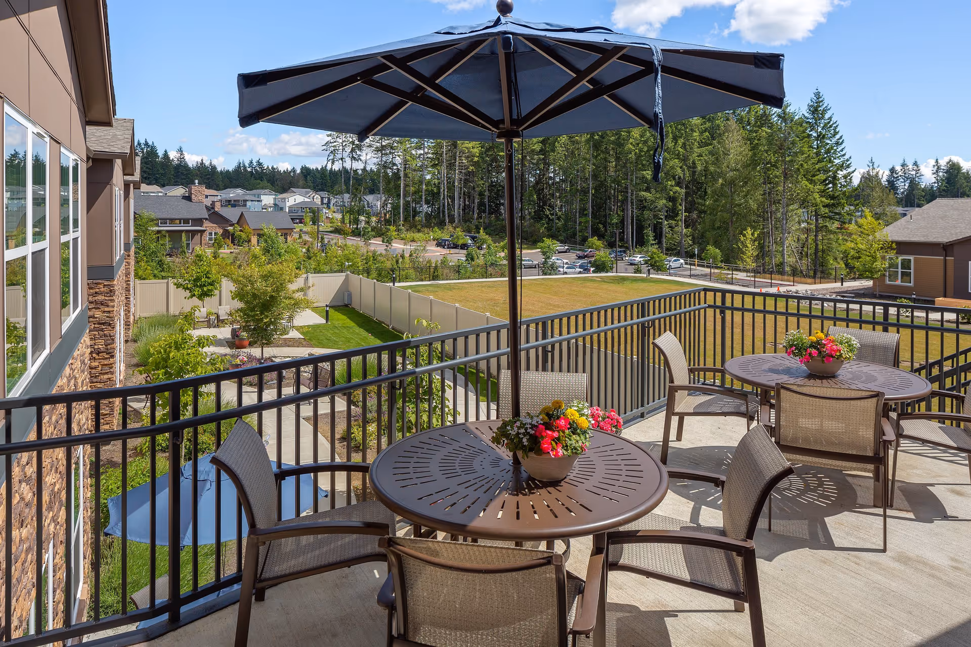 Outdoor patio area at Heron's Key with round metal tables and chairs, each table decorated with a flower pot. Large blue umbrellas provide shade. The patio overlooks a fenced grassy area, trees, and a parking lot with cars under a partly cloudy sky.