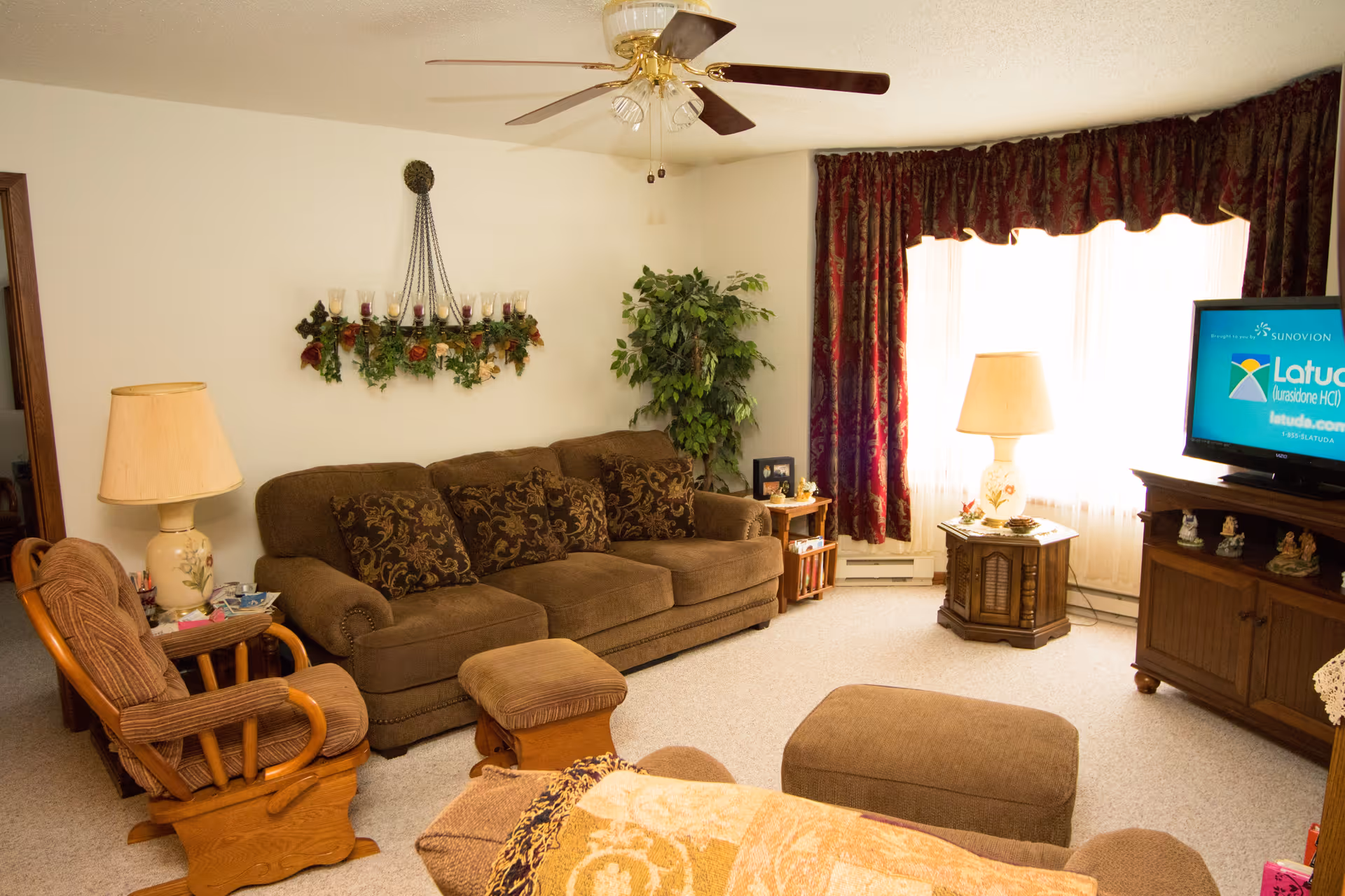 Cozy living room with a brown sofa, armchairs and ottomans, a TV on a wooden stand, side tables with lamps, and a window with curtains.