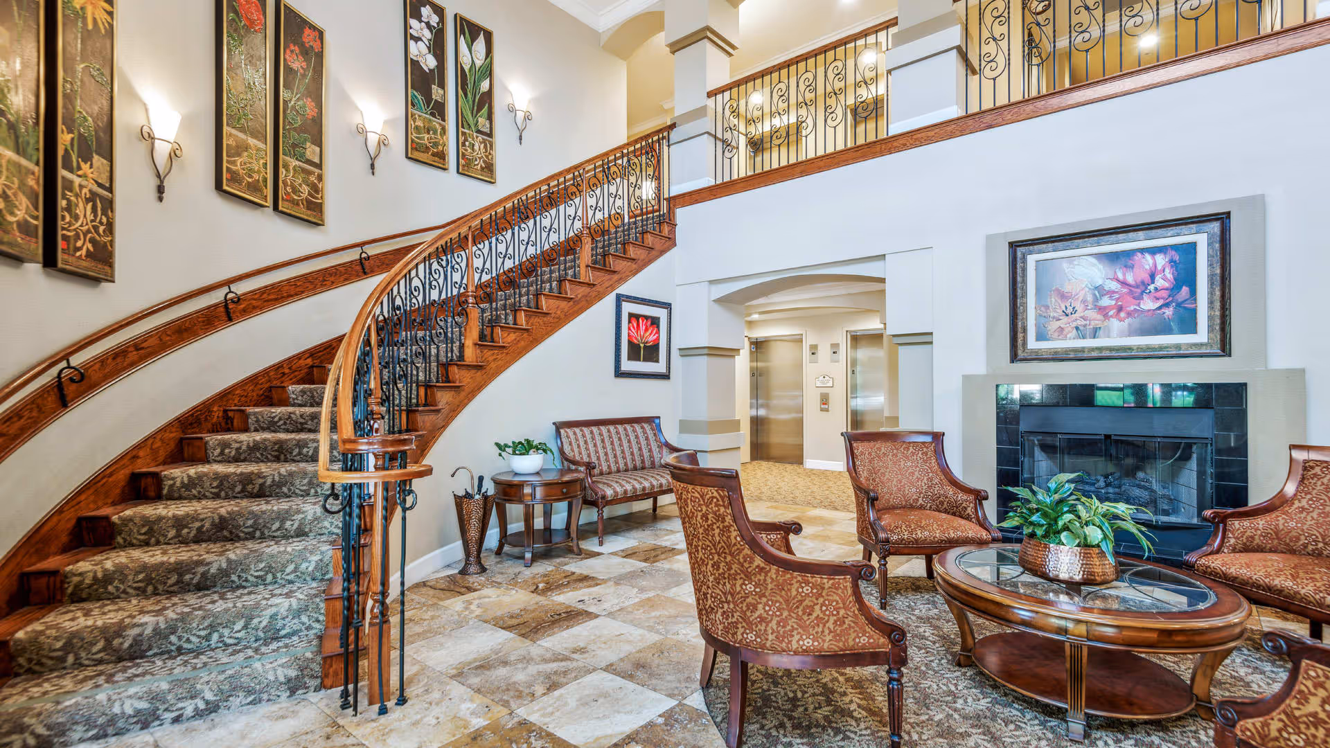 A spacious and elegant interior common area featuring a curved wooden staircase with ornate iron railings, a fireplace with a floral painting above it, and a seating arrangement including patterned upholstered chairs and a glass-top coffee table with a plant. The walls are decorated with framed botanical artwork and sconces, and there are elevators visible in the background.