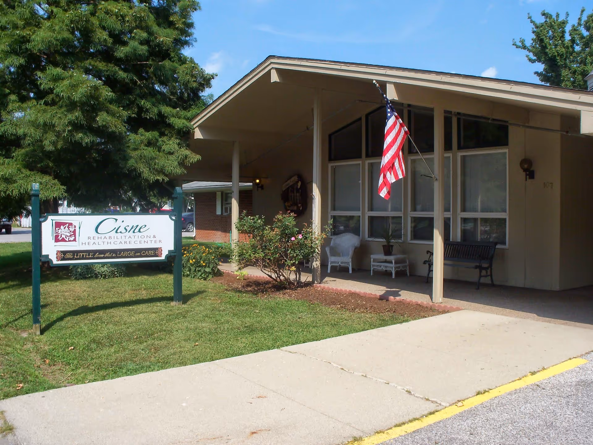 Exterior view of Cisne Rehabilitation and Health Care Center building with a covered porch, American flag, benches, and a sign on the lawn displaying the facility name.