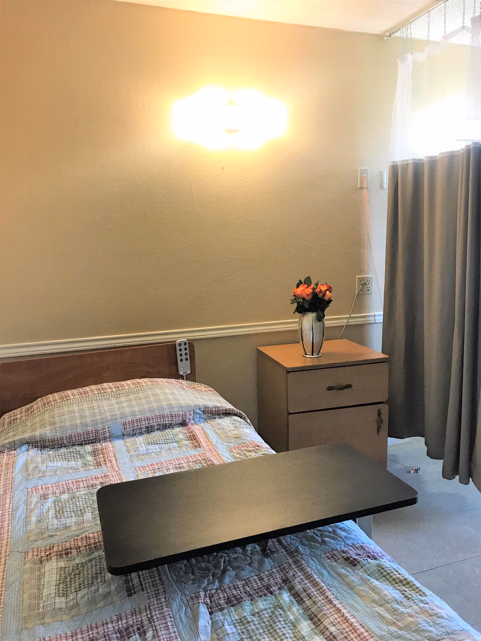 A single bed with a patterned quilt and a black overbed table in a room. Next to the bed is a wooden nightstand with a vase of orange flowers. A wall-mounted light fixture is above the bed, and a window with sheer and dark curtains is visible on the right side.