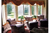 Sunlit sitting area with patterned armchairs and small tables arranged in front of large windows with potted plants.