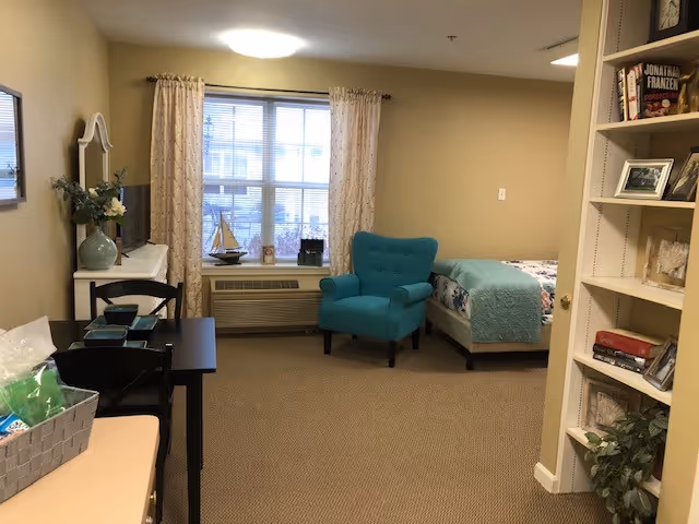 Studio-style senior living room with a bed, teal armchair, window with curtains, small desk, TV and bookshelf.