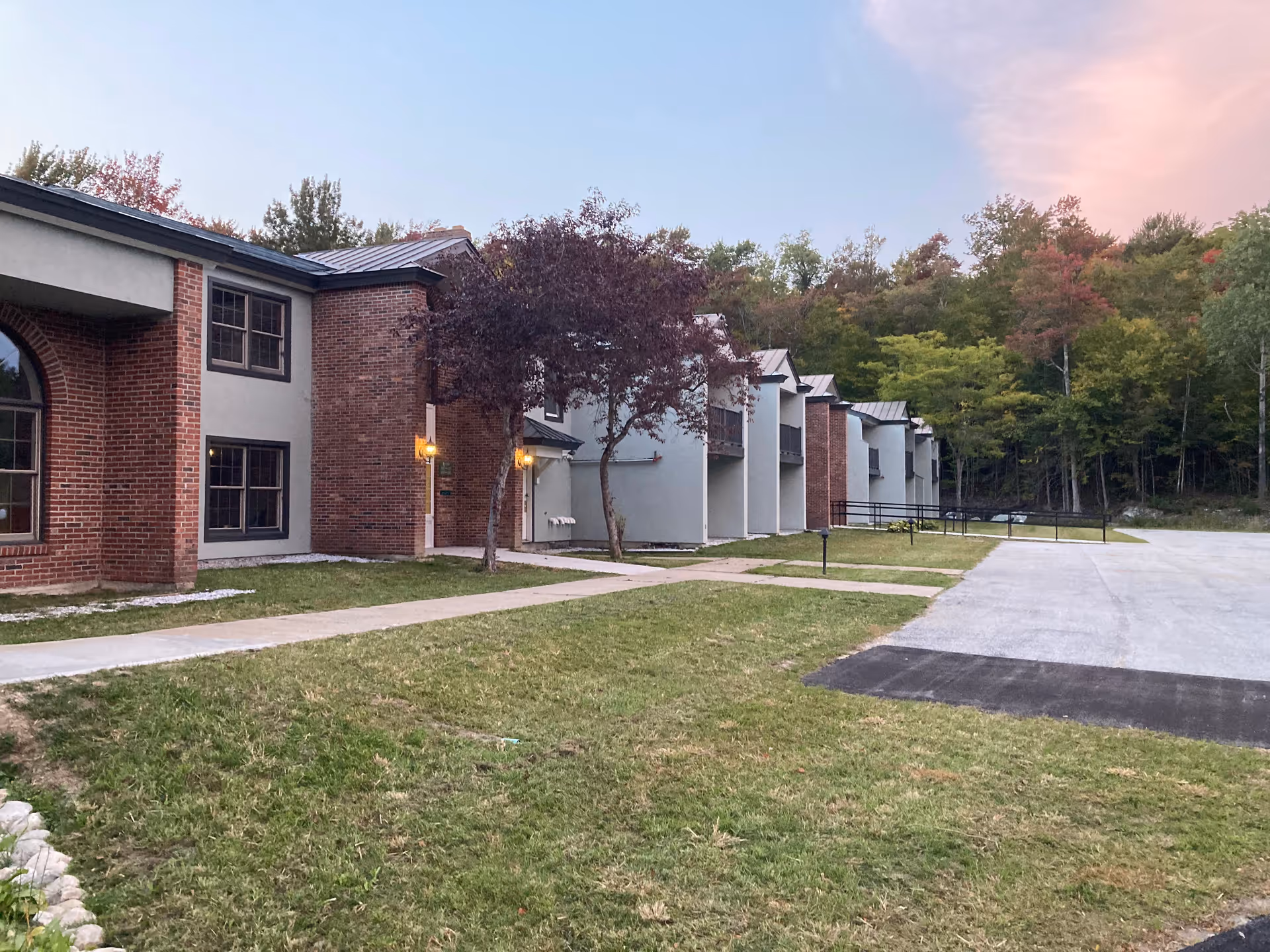 Exterior view of a senior living facility building with brick and light gray walls, multiple windows, and small trees in front. There is a paved walkway and a parking area adjacent to the building, with a backdrop of trees and a partly cloudy sky at sunset.