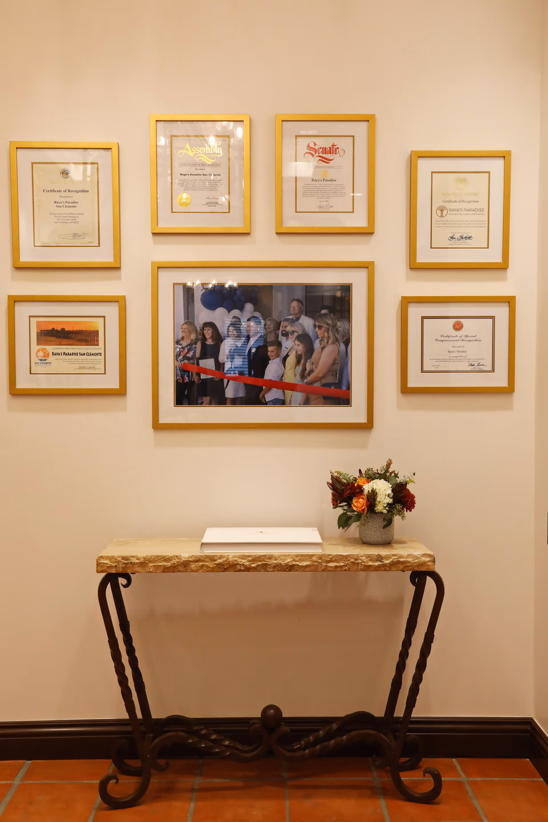 A wall display featuring seven framed certificates and a central framed photo of a ribbon-cutting ceremony with a group of people. Below the frames is a decorative table with a stone top and wrought iron legs, holding a closed book and a small flower arrangement in a vase. The setting appears to be an interior hallway or lobby area.
