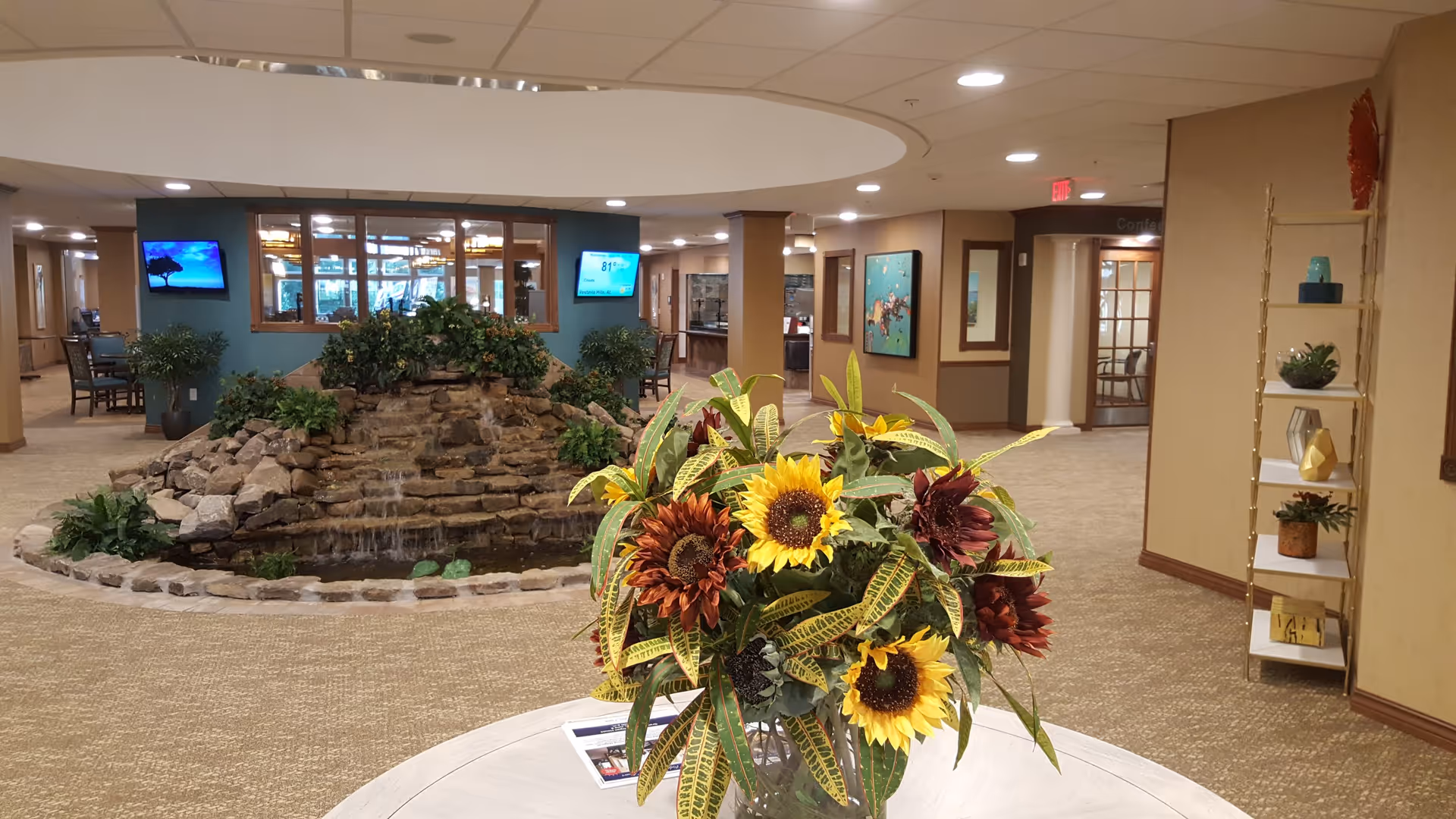 Interior view of a retirement community common area featuring a round table with a large bouquet of sunflowers and other flowers in the foreground. Behind the table is a decorative rock water feature with plants. The space has beige walls, carpeted floors, and recessed lighting. There are two wall-mounted TVs, framed artwork, and a shelving unit with decorative items on the right side.