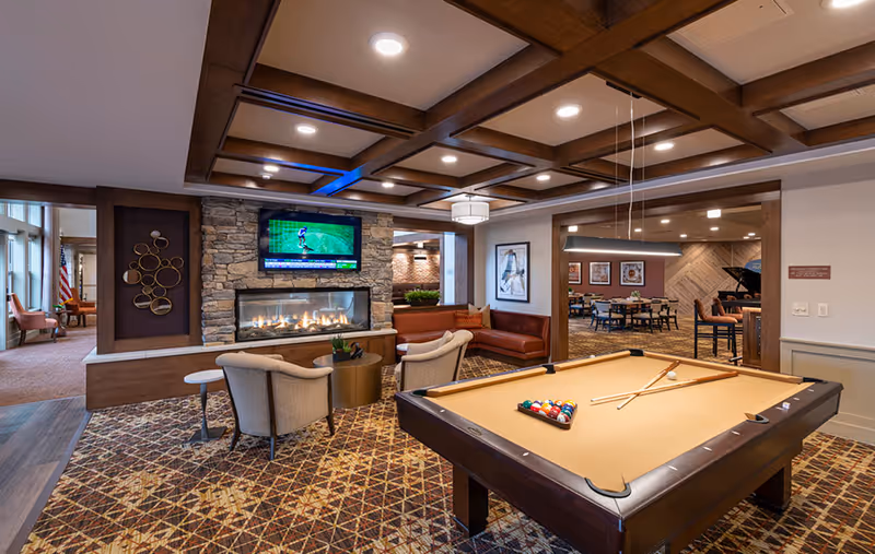 A cozy senior living common room with a pool table in the foreground, seating around a stone fireplace with a mounted TV, and a dining area beyond.