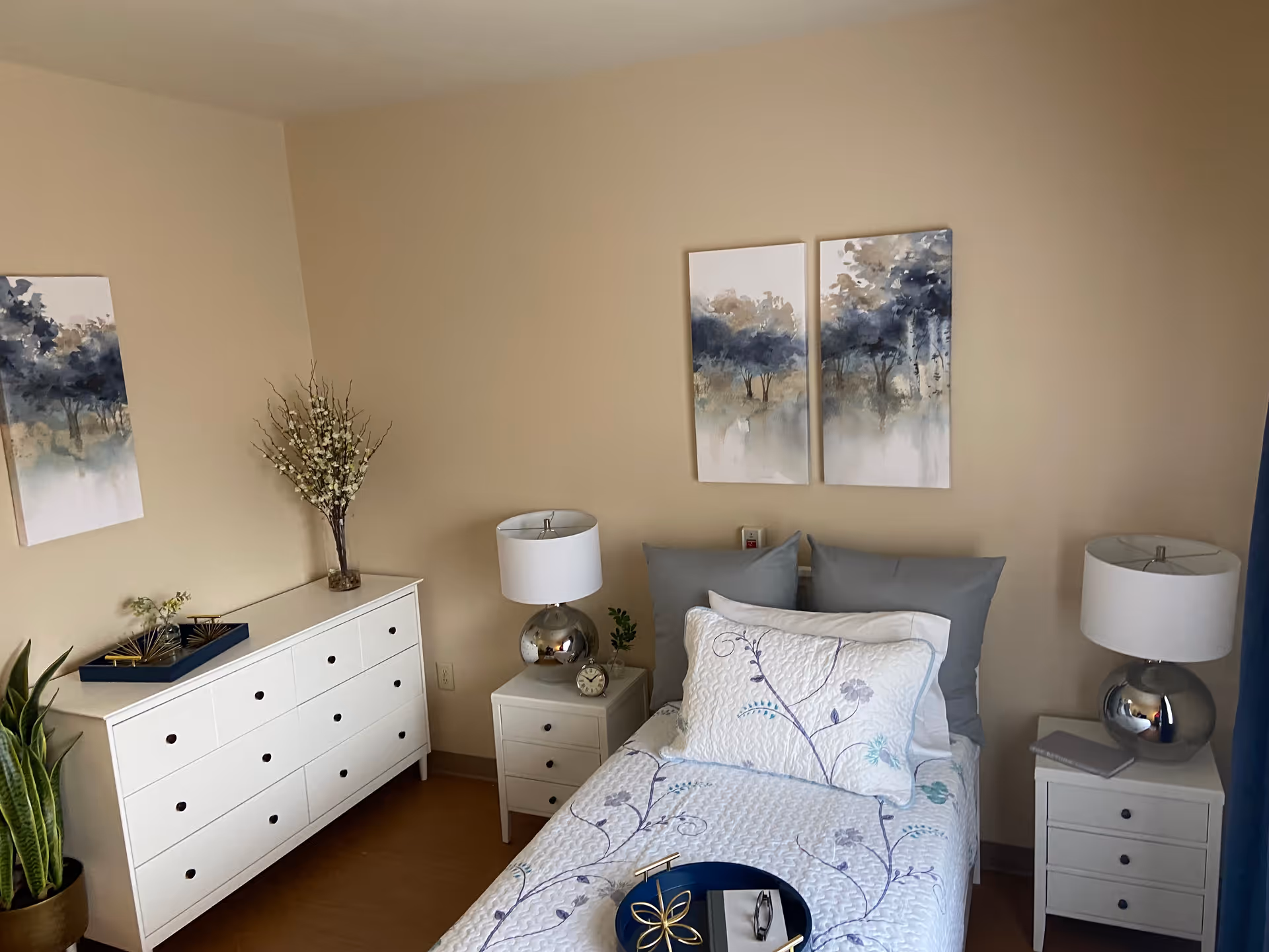 A neatly arranged bedroom with a single bed featuring white and floral bedding, two gray pillows, and a decorative tray on the bed. On either side of the bed are white nightstands with silver lamps. A white dresser with multiple drawers is positioned against the wall, decorated with a vase of flowers and a tray with decorative items. The walls are beige and adorned with three abstract paintings depicting trees in blue and beige tones. A green potted plant is placed on the floor next to the dresser.