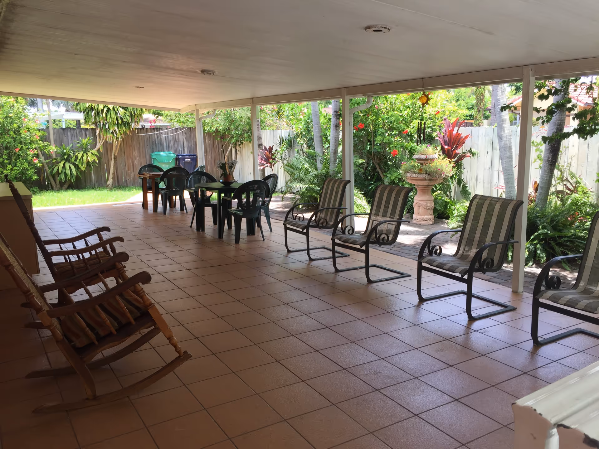 Covered outdoor patio area with tiled floor, several chairs including rocking chairs and metal chairs with cushions, small tables with plastic chairs, surrounded by lush green plants and flowers, with a wooden fence in the background.