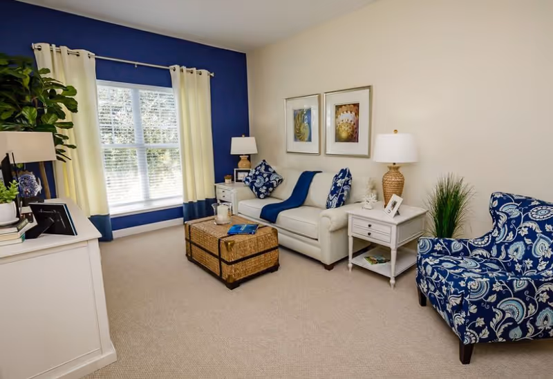 Bright living room with a white sofa, blue patterned armchair, wicker coffee table, and a large window with cream curtains against a blue accent wall.