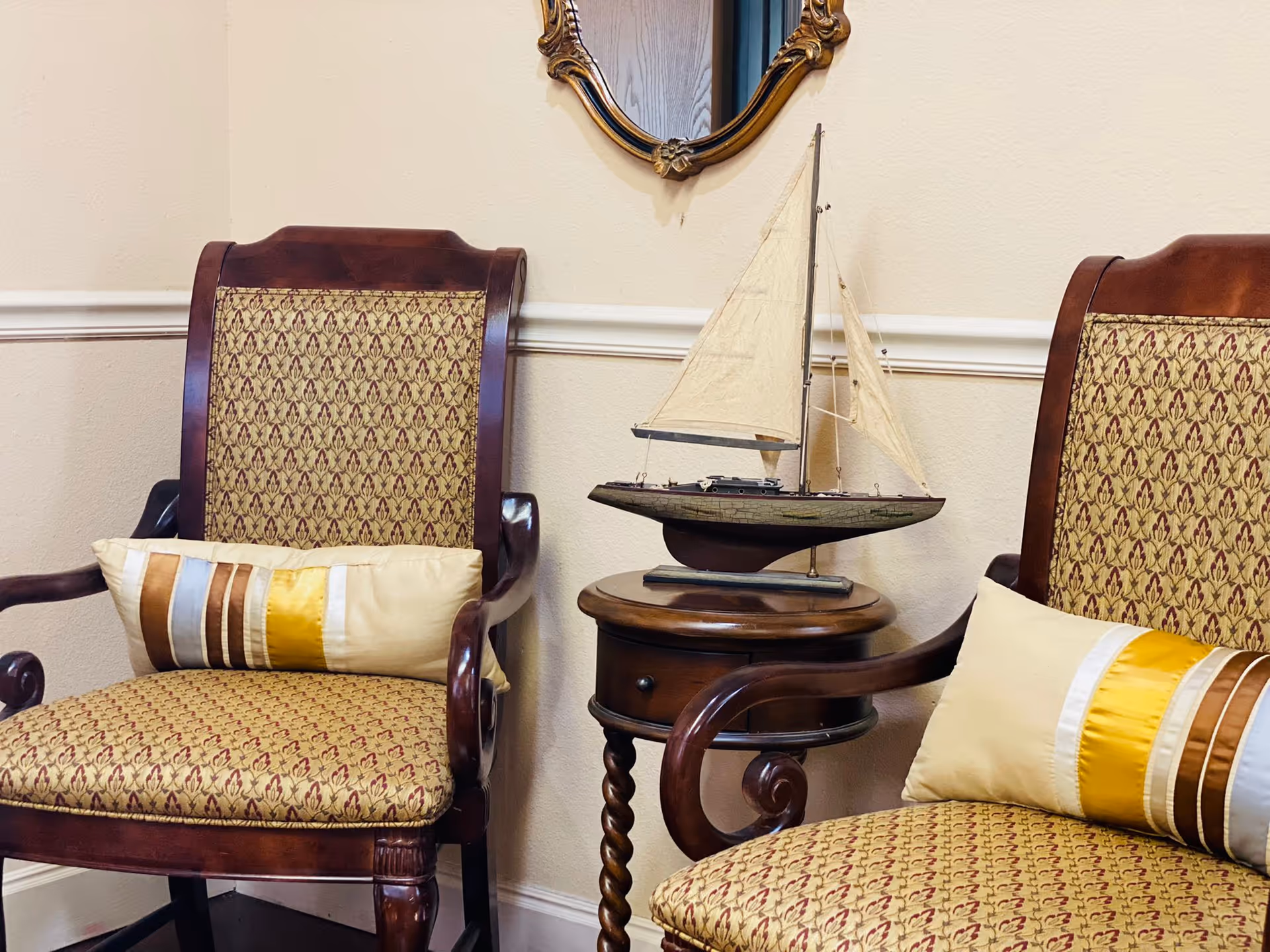Two wooden armchairs with patterned upholstery and decorative pillows are placed on either side of a small round wooden table. On the table is a model sailboat. A decorative mirror with a gold frame hangs on the wall above the table. The setting appears to be a cozy interior corner.