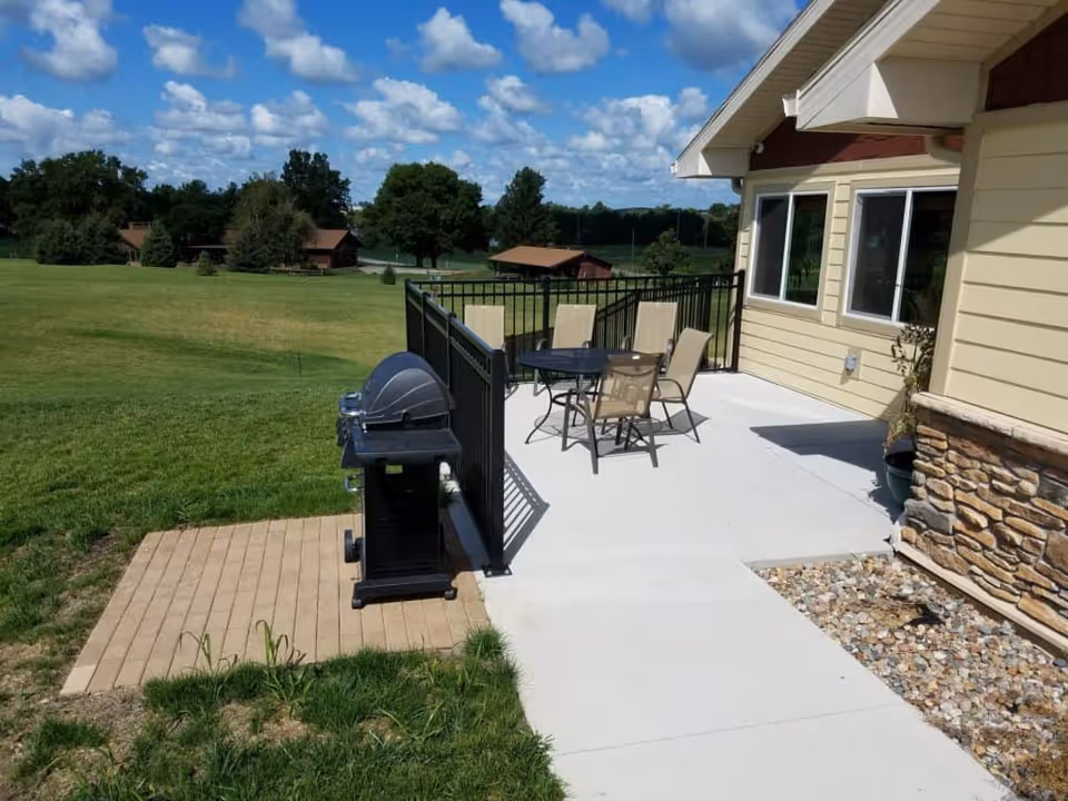 Outdoor patio area at a senior living facility with a black metal table and four beige chairs on a concrete surface. A black grill is positioned on a small paved section next to the patio. The building exterior features beige siding with stone accents. In the background, there is a large grassy field with trees and small wooden structures under a partly cloudy blue sky.