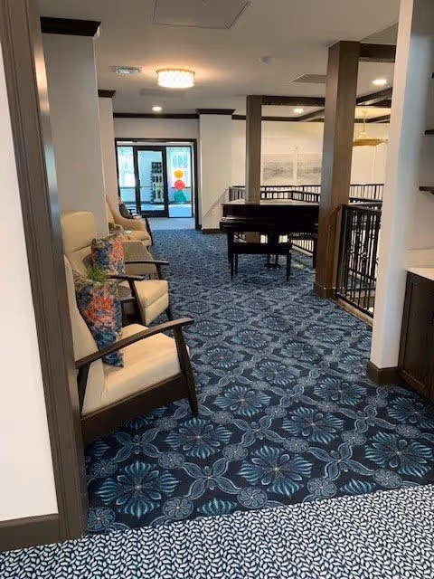 Interior common area with patterned blue carpet, chairs with floral pillows along the left, a piano and railing in the center, and glass doors at the far end.