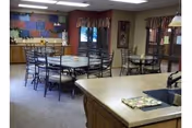 Interior view of a dining area in a retirement community featuring multiple round tables with chairs, a countertop with a sink in the foreground, and colorful artwork on the far wall. The room has windows with valance curtains and neutral-colored walls and carpet.