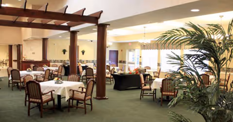 A spacious dining room with multiple tables covered in white tablecloths, each surrounded by wooden chairs with cushions. The room features large windows allowing natural light, green carpeting, wooden columns, and decorative plants.