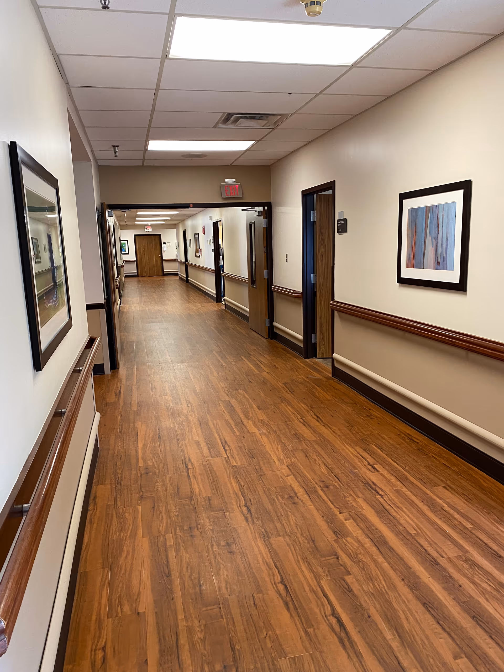A long, clean hallway with wood-patterned flooring and beige walls in a healthcare facility. The hallway has handrails on both sides, framed artwork on the walls, and several open doors leading to rooms. The ceiling has fluorescent lighting and an exit sign is visible at the far end.