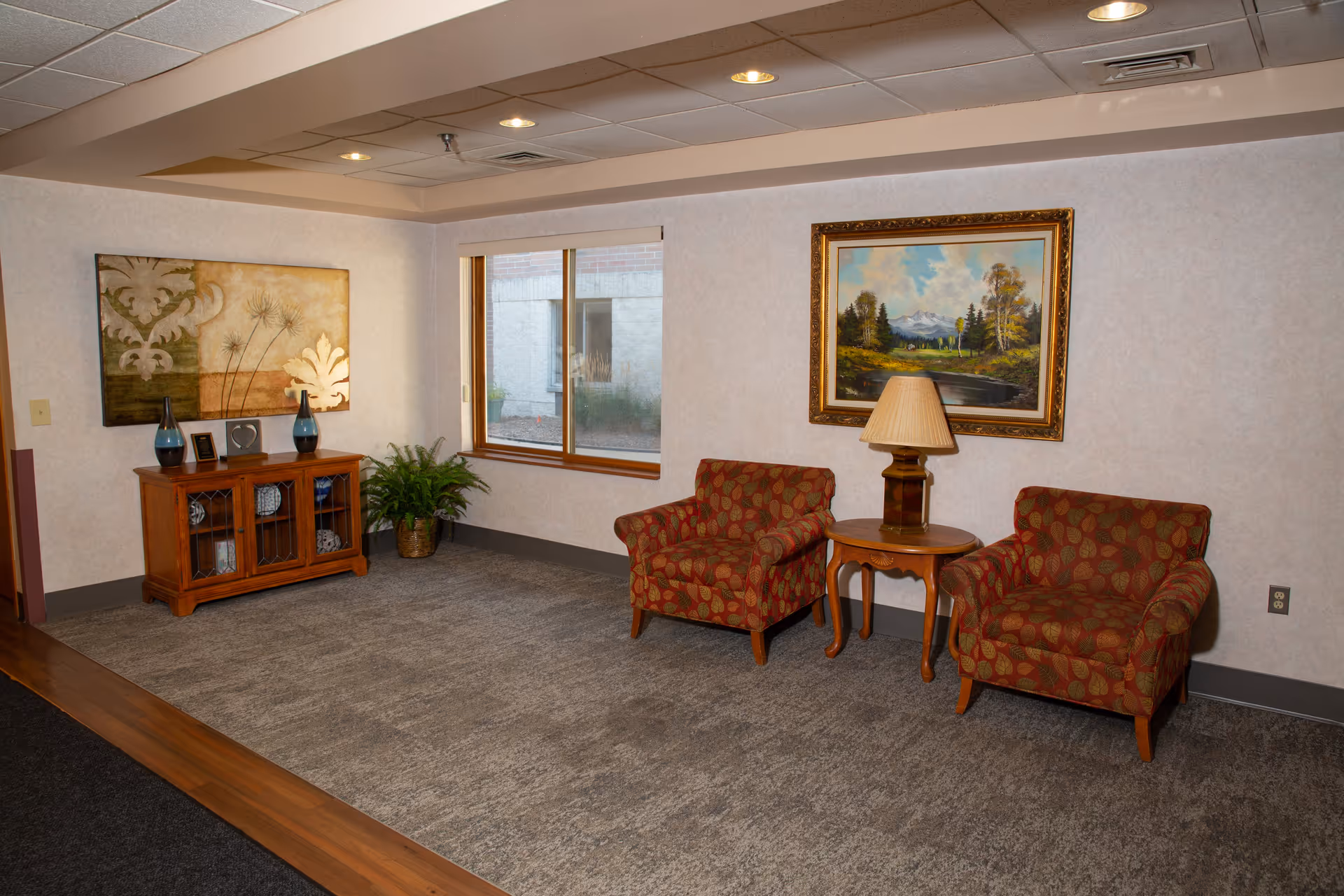 A cozy sitting area in a senior living facility with two patterned armchairs separated by a wooden side table with a lamp. Behind the chairs is a framed landscape painting. To the left, there is a wooden cabinet with decorative items and two blue vases on top, a potted plant next to it, and a window showing an exterior wall outside.